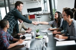  A meeting room full of people sitting at a table having a discussion about onboarding whilst one man stands and shakes hand with a female employee