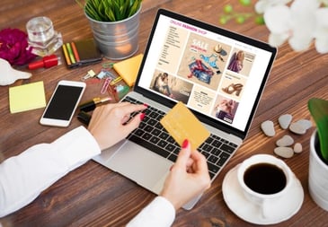 A women at a desk surrounded by a coffee and her phone whilst on a laptop holding her credit card in one hand and online shopping for fast fashion
