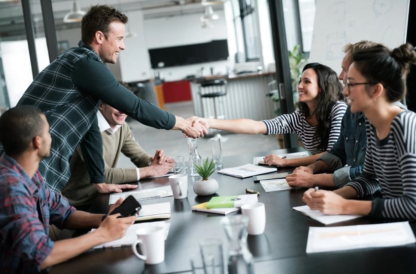 A meeting room full of people sitting at a table having a discussion about onboarding whilst one man stands and shakes hand with a female employee