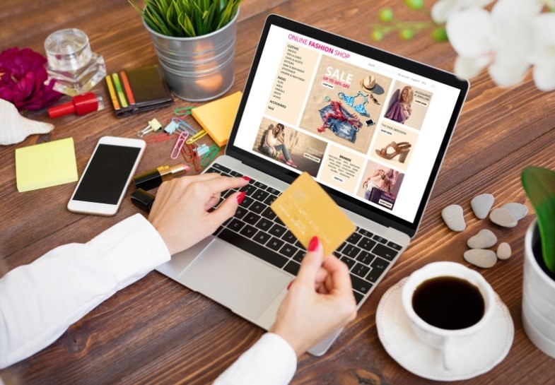 A women at a desk surrounded by a coffee and her phone whilst on a laptop holding her credit card in one hand and online shopping for fast fashion